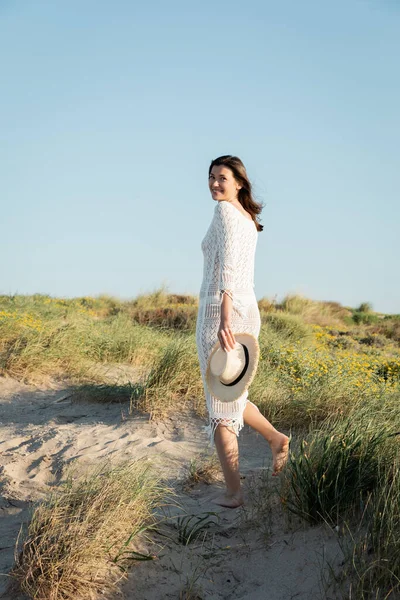 Mujer joven sosteniendo sombrero de sol mientras camina sobre hierba en la playa - foto de stock