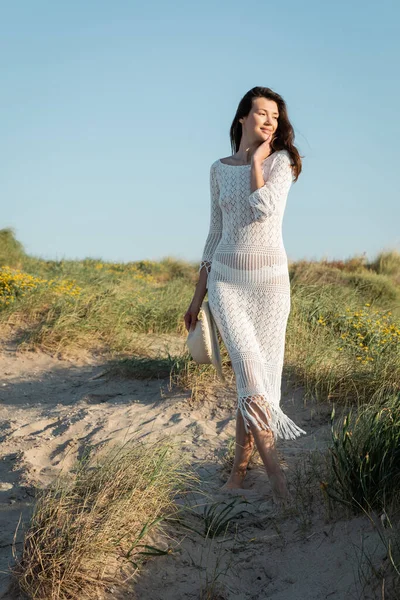 Barefoot woman in knitted dress holding sun hat while standing on beach — Stock Photo