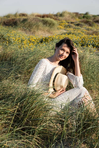 Mujer morena en vestido de verano sosteniendo sombrero de paja mientras está sentado en la hierba en la playa - foto de stock