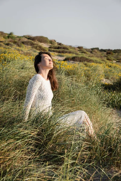 Brunette woman closing eyes while sitting on grass on beach — Stock Photo