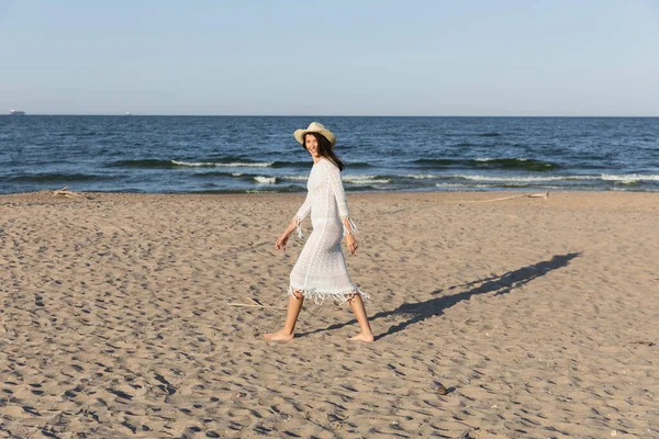 Donna felice in abito e cappello da sole guardando la fotocamera mentre cammina sulla spiaggia vicino al mare — Foto stock