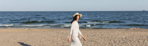Mujer positiva en vestido y sombrero de paja caminando en la playa cerca del mar, bandera - foto de stock