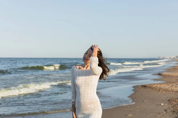 Brunette woman in knitted dress touching hair near sea on beach — Stock Photo