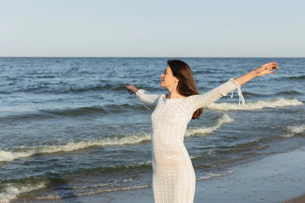 Vista lateral de mujer morena joven en vestido de punto mirando el mar en la playa - foto de stock