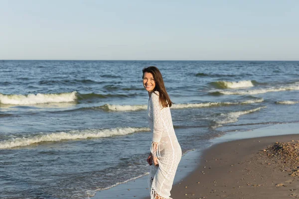 Jolie femme en robe souriant à la caméra près de la mer sur la plage — Photo de stock