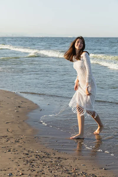 Positive young woman in knitted dress standing on wet sand on beach — Stock Photo