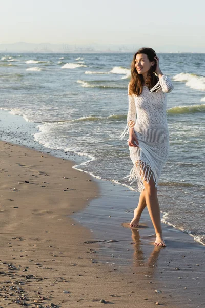 Positive young woman in dress walking on wet sand near sea — Stock Photo
