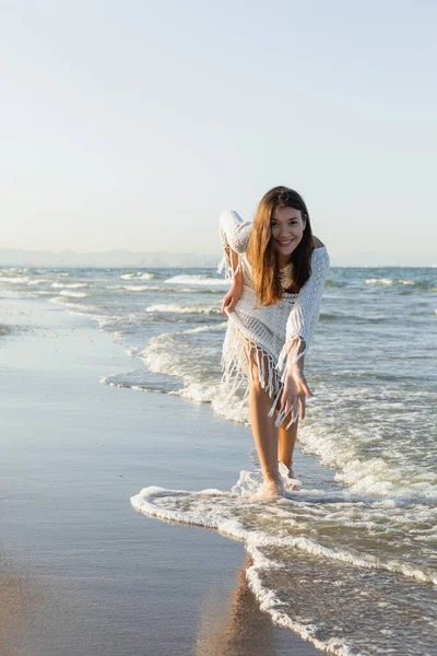 Happy woman in dress looking at camera while standing in sea water on sandy beach — Stock Photo