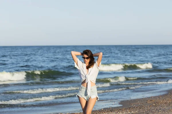 Young woman in sunglasses and denim shorts standing near blurred sea — Stock Photo
