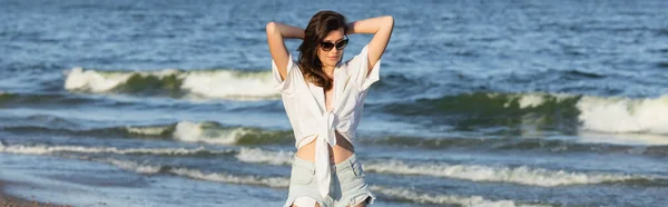 Brunette femme en lunettes de soleil et chemise blanche debout près de la mer floue, bannière — Photo de stock