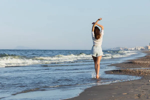 Vista posteriore della donna in pantaloncini di jeans e camicia che cammina vicino al mare sulla spiaggia — Foto stock