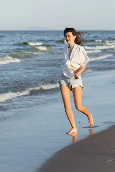 Mujer joven en camisa y pantalones cortos corriendo cerca del mar en la playa - foto de stock