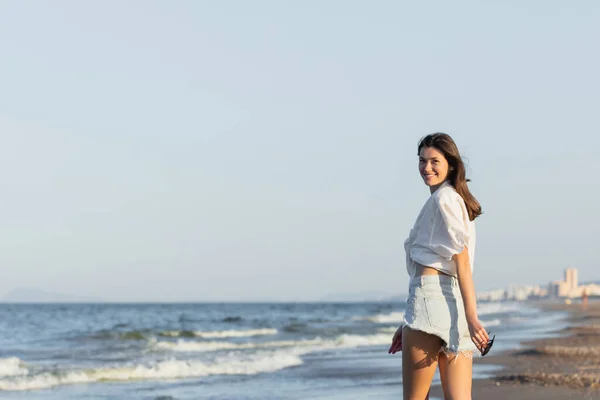 Happy young woman holding sunglasses and looking at camera near sea — Stock Photo