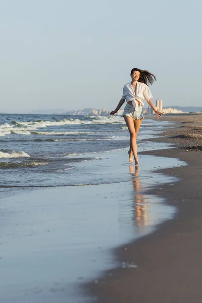 Mujer morena alegre en pantalones cortos sosteniendo gafas de sol mientras corre en la playa - foto de stock