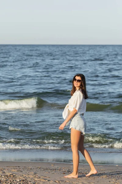 Positive junge Frau mit Sonnenbrille spaziert auf nassem Sand am Strand — Stockfoto