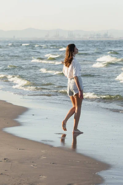 Vista laterale di giovane donna in pantaloncini in piedi su sabbia bagnata vicino al mare — Foto stock