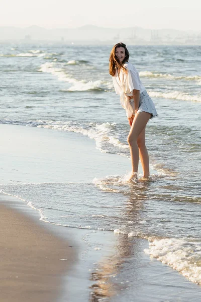 Sorridente donna in camicia guardando la fotocamera mentre in piedi in mare — Foto stock