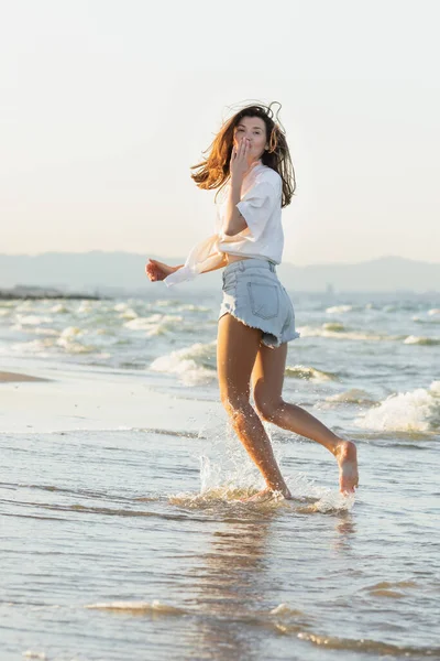 Молода жінка дме повітряний поцілунок під час бігу на морському узбережжі — стокове фото