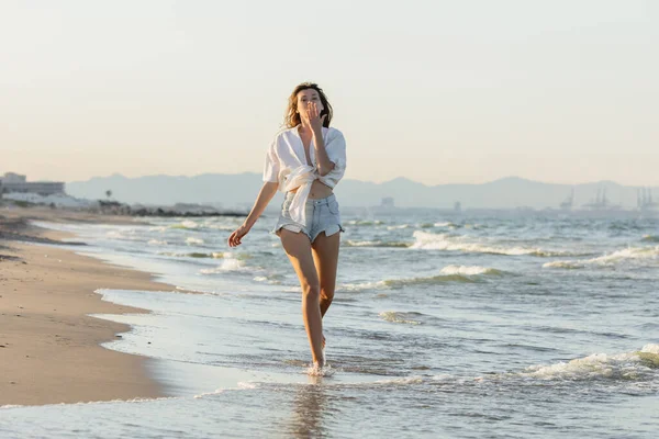 Brunette femme en chemise et short soufflant l'air baiser tout en marchant sur la côte de la mer — Photo de stock