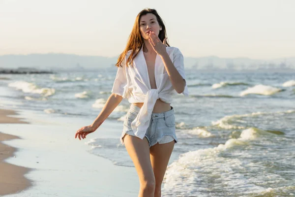 Молодая женщина дует воздушный поцелуй перед камерой во время прогулки по пляжу у моря — стоковое фото