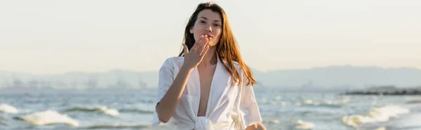 Bruna donna in camicia soffiando aria bacio con il mare sullo sfondo, banner — Foto stock