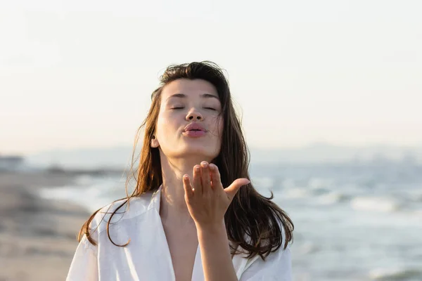 Brunette woman in white shirt blowing air kiss on blurred beach — Stock Photo