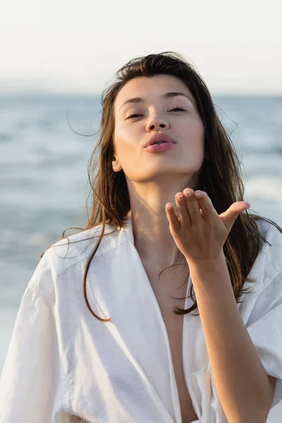 Brunette woman in shirt blowing air kiss near blurred sea — Stock Photo