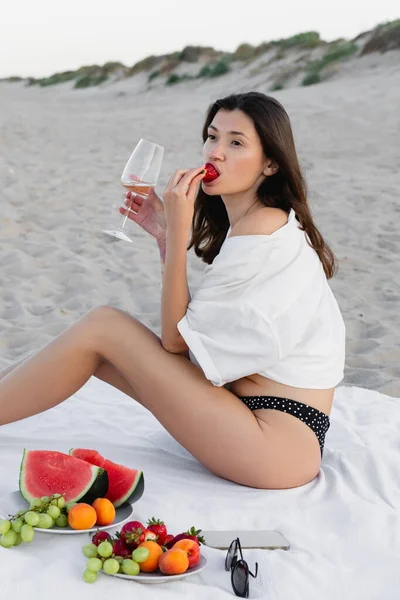 Жінка в сорочці їсть полуницю і тримає вино біля смартфона і сонцезахисних окулярів на пляжі. — стокове фото