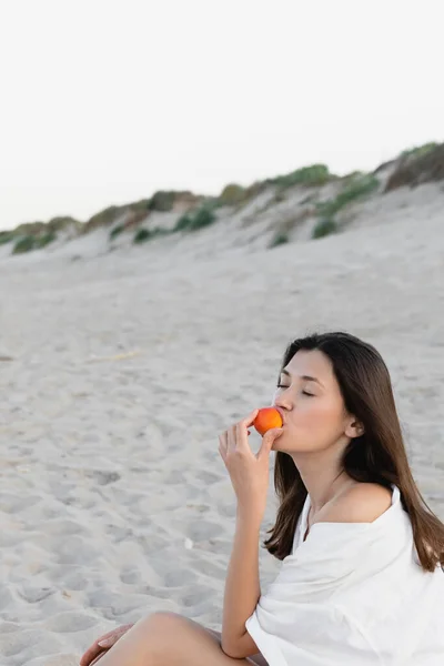 Mujer morena en camisa comiendo albaricoque en la playa - foto de stock