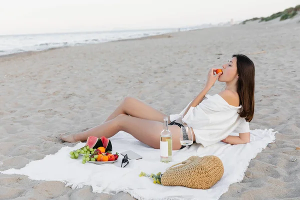 Vista lateral de la mujer en traje de baño y camisa comiendo albaricoque cerca del vino y el teléfono inteligente en la playa - foto de stock