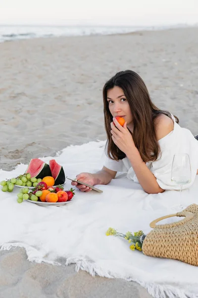 Mujer joven en camisa comiendo albaricoque y usando teléfono inteligente cerca de frutas y vino en la playa - foto de stock