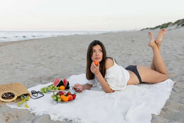 Mujer morena en traje de baño y camisa sosteniendo teléfono inteligente y fruta cerca del vino en manta en la playa - foto de stock