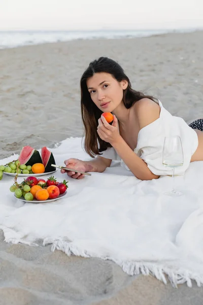 Woman in shirt holding apricot and smartphone near wine on blanket on beach — Stock Photo