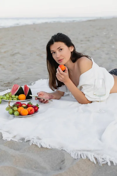 Mulher de camisa segurando damasco e smartphone perto de vinho em cobertor na praia — Fotografia de Stock