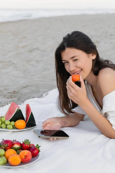 Jovem mulher de camisa segurando damasco e usando smartphone perto de frutas na praia — Fotografia de Stock