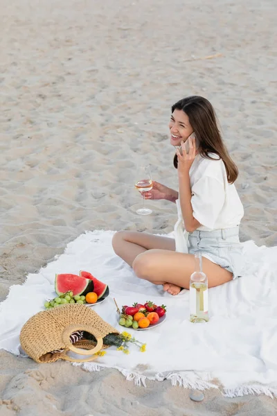 Smiling woman talking on smartphone and holding wine near fruits on beach — Stock Photo