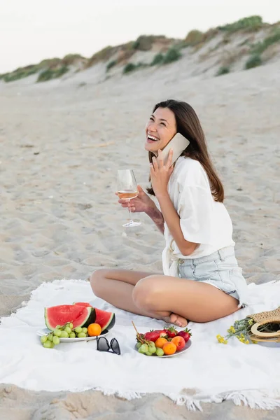 Donna positiva che parla sullo smartphone e tiene il vino vicino alla frutta fresca sulla coperta sulla spiaggia — Foto stock