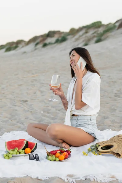 Mujer sonriente hablando en smartphone y sosteniendo vino cerca de frutas maduras en manta en la playa - foto de stock