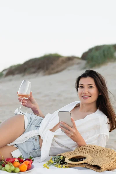 Mujer sonriente usando smartphone y sosteniendo copa de vino cerca de frutas y bolso en la playa - foto de stock