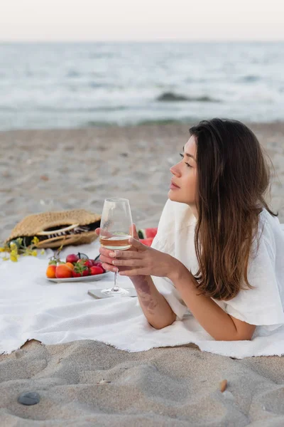 Vista laterale della giovane donna che tiene il bicchiere di vino vicino ai frutti sfocati sulla spiaggia — Foto stock