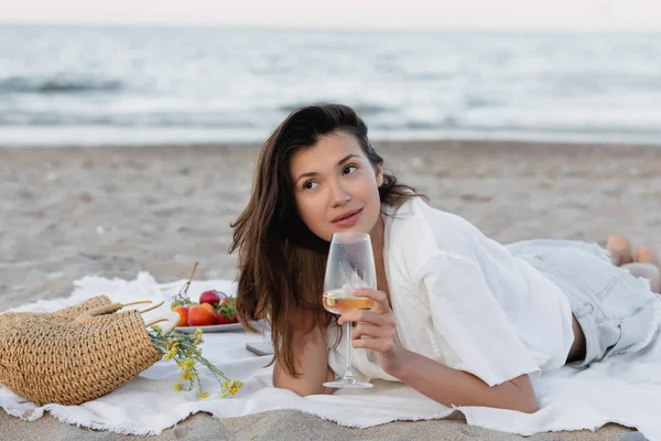 Mujer morena sosteniendo copa de vino cerca de bolso y frutas en manta en la playa - foto de stock