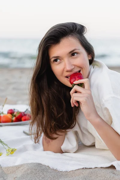 Mujer morena sonriente comiendo fresa en manta en la playa - foto de stock