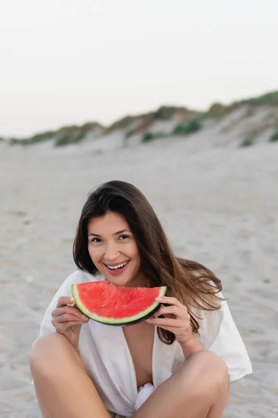 Mujer alegre en camisa sosteniendo sandía madura en la playa - foto de stock