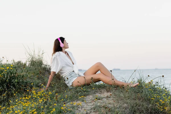 Seitenansicht einer Frau mit Kopfhörern und Hemd, die abends am Strand sitzt — Stockfoto