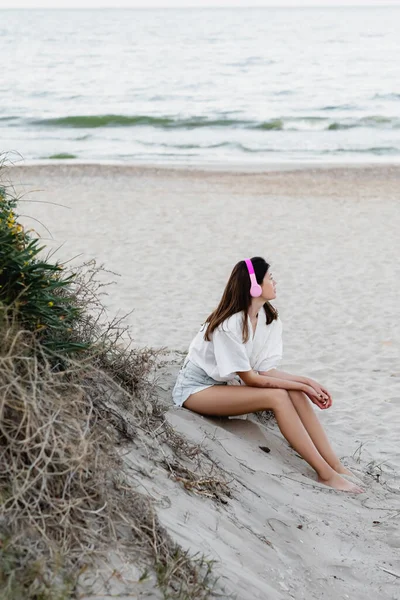 Vista lateral de mujer joven en camisa escuchando música en auriculares en la playa cerca del mar - foto de stock