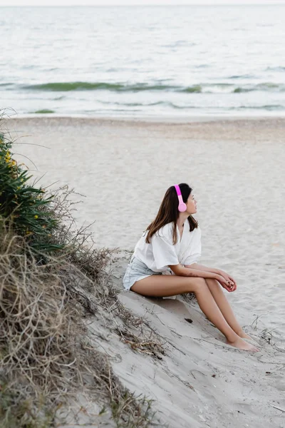 Side view of young woman in shirt listening music in headphones on beach near sea — Stock Photo