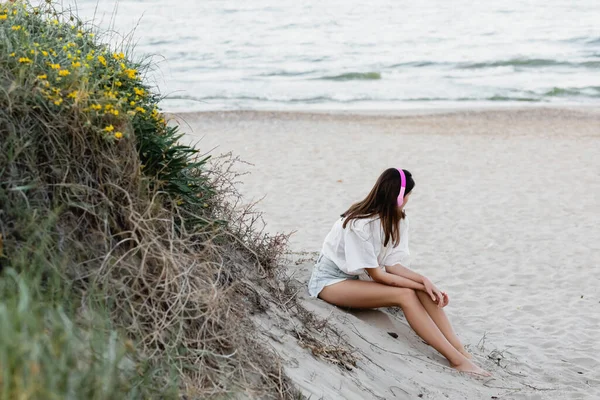 Молодая женщина в наушниках сидит на песке у моря — стоковое фото