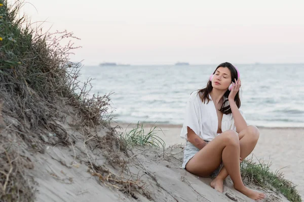 Mujer con auriculares cerrando los ojos mientras está sentada en la playa por la noche - foto de stock