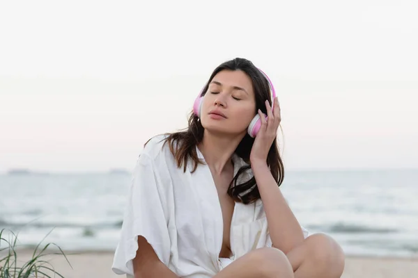 Young woman in shirt and headphones sitting on beach near blurred sea — Stock Photo