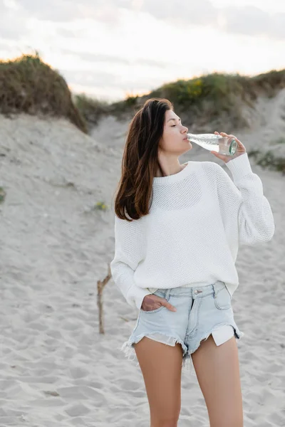 Seitenansicht einer jungen Frau, die am Strand Mineralwasser trinkt — Stockfoto